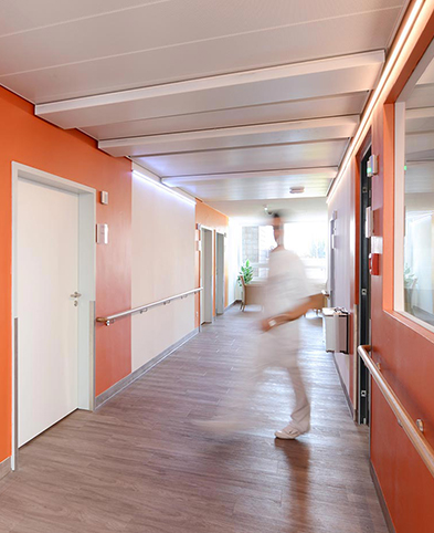 Pflegefachkraft kommt aus dem Dienstzimmer der geriatrischen Klinik – Flurwände sind in verschiedenen Orangetönen gestrichen.