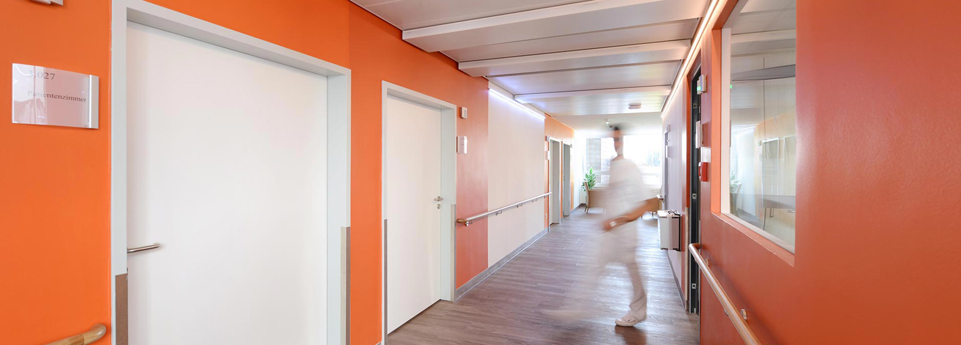Pflegefachkraft kommt aus dem Dienstzimmer der geriatrischen Klinik – Flurwände sind in verschiedenen Orangetönen gestrichen.