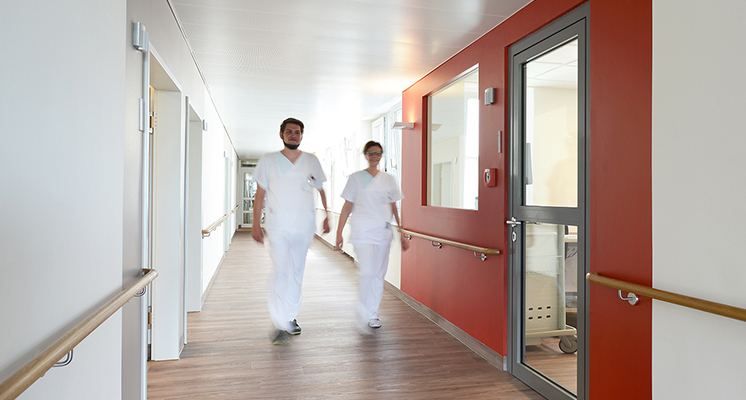 Zwei Pflegefachkräfte laufen am Dienstzimmer und mehreren Patientenzimmern der Palliativstation vorbei.