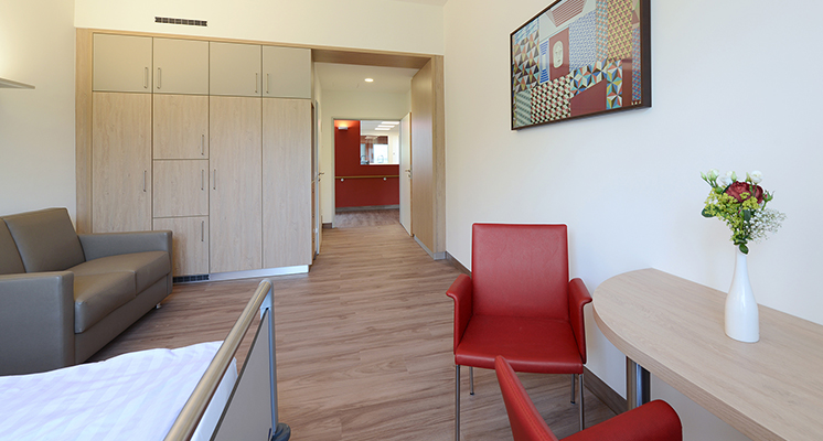 Patientenzimmer der Palliativstation inkl. Schlafsofa für Familienmitglieder und Freunde, Sitzbereich und Schrankwand in Holzoptik.