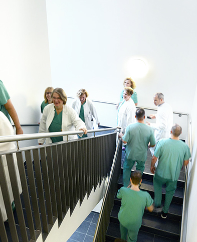 Eine Gruppe vom Mitarbeitern mit Kasaks und Kitteln bekleidet gehen im Treppenhaus eine Etage hoch.