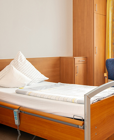 Zimmer der Kurzzeitpflege – freundlich eingerichtet mit funktionalem Bett, Sitzbereich am Fenster und verschiedenen Schränken.