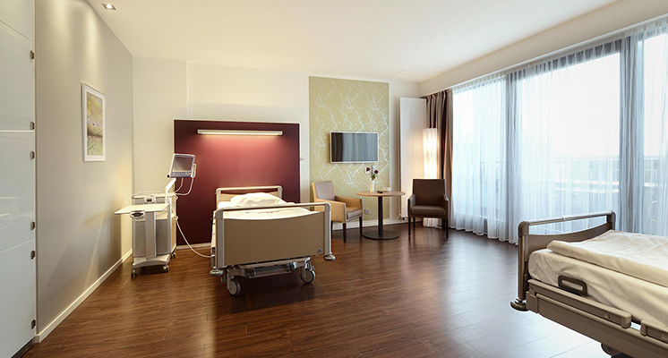Komfortables 2-Bett-Zimmer der Station 4E mit Sitzbereich, hochwertiger Wandgestaltung und Nachttisch inkl. Entertainment-System.