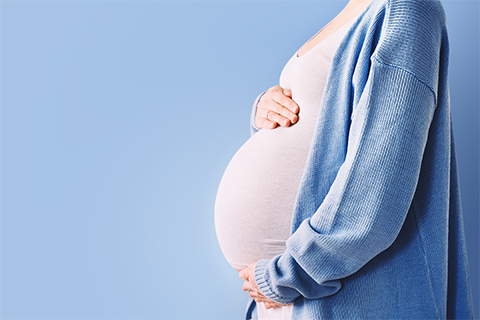 Schwangere hält ihre Hände oben und unten an ihren Bauch – nur dieser Ausschnitt ist vor einem hellblauen Hintergrund zu sehen. Die Frau trägt eine hellblaue Strickjacke und ein weißes Shirt.