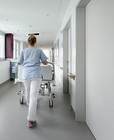 Man sieht eine Pflegefrachkraft von hinten, die ein Patientenbett über einen Stationsflur schiebt.