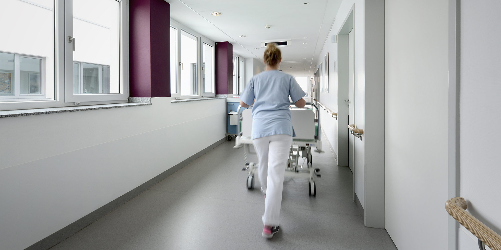 Man sieht eine Pflegefrachkraft von hinten, die ein Patientenbett über einen Stationsflur schiebt.