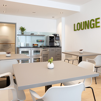 Lounge der Komfortstation 3E – mit hellen Stühlen, grauen Tischen, einer Küchenzeile mit Edelstahlfronten und einem Moos-Schriftzug 