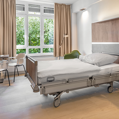 Patientenzimmer auf der Komfortstation 3E mit Blick in Richtung Fenster – im Vordergrund das Patientenbett und dahinter eine Sitzgruppe am Fenster sowie eine Leseecke bestehend aus Stuhl und Stehlampe.